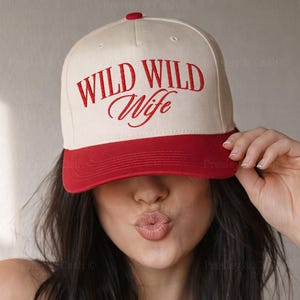 May include: A baseball cap with a cream-colored crown and a red brim. The cap features the words "WILD WILD Wife" in red script across the front. The hat is being held up by a person.
