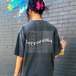 K&ouml;nnte beinhalten: Ein schwarzes Oversize-T-Shirt mit dem Text "Let's Go Girls", der sechsmal in Wei&szlig; wiederholt wird. Das Model tr&auml;gt das Shirt und wei&szlig;e Sneaker. Der Text "Model is normally a MEDIUM but sized up to an XXL for this look!" ist am unteren Rand des Bildes sichtbar.