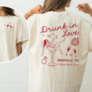 May include: Ivory t-shirt with a dark red design. The design features a woman in a martini glass with the text "Drunk in Love", a disco ball, and the words "Nashville, TN Kate's Back Trip".