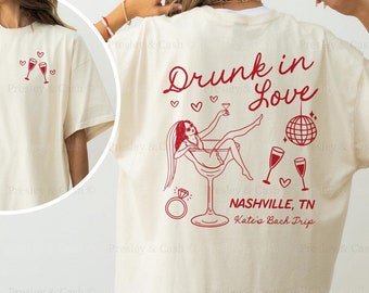 May include: Ivory t-shirt with a dark red design. The design features a woman in a martini glass with the text "Drunk in Love", a disco ball, and the words "Nashville, TN Kate's Back Trip".