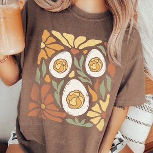 May include: A brown t-shirt with a graphic design of three eggs surrounded by colorful flowers. The text "ESPRESSO-L" is printed on the bottom of the shirt.