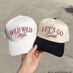 May include: Two baseball caps, one white and one natural, with maroon embroidery. The white cap reads "WILD WILD Wife," while the natural cap says "LET'S GO Girls." The natural cap has a black brim.