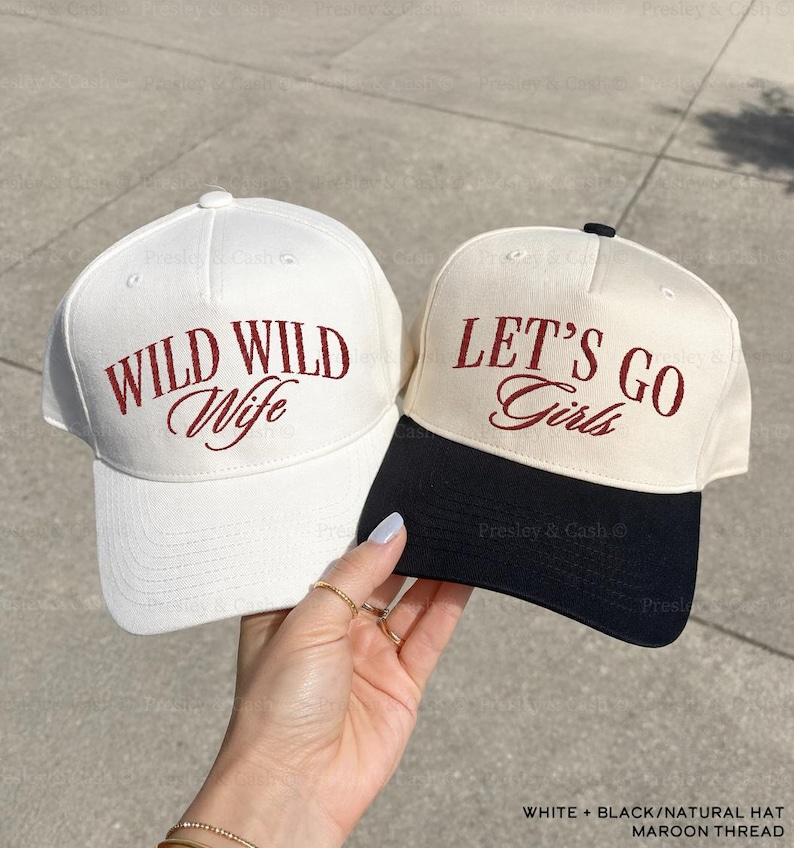 May include: Two baseball caps, one white and one natural, with maroon embroidery. The white cap reads "WILD WILD Wife," while the natural cap says "LET'S GO Girls." The natural cap has a black brim.