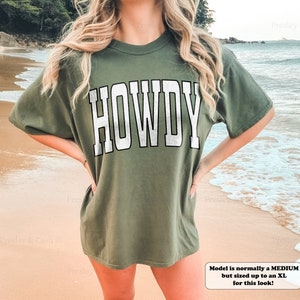 May include: A green t-shirt with the word "HOWDY" printed in white on the front. The model is wearing a size XL.