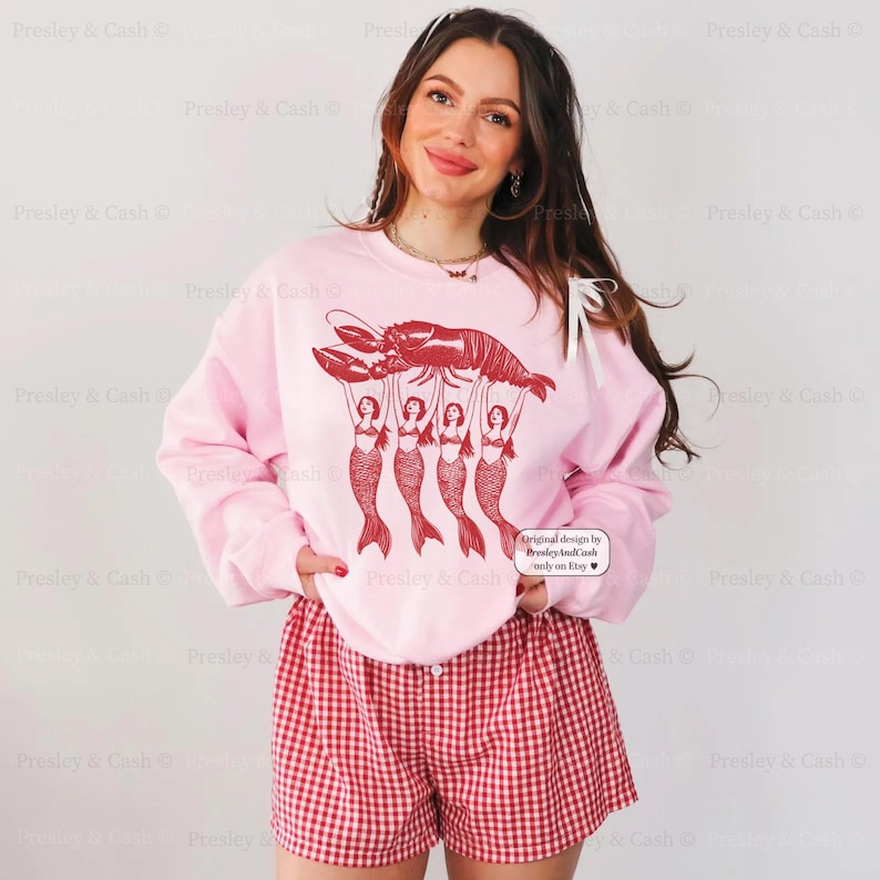 May include: Pink sweatshirt with a red lobster graphic held up by four mermaids. The sweatshirt is paired with red and white checkered shorts. The image includes the text "Original design by Presley & Cash only on Etsy."