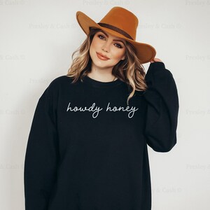 Pode incluir: Uma camisa de moletom preta com o texto "howdy honey" em escrita cursiva branca. A modelo est&aacute; usando um chap&eacute;u marrom.