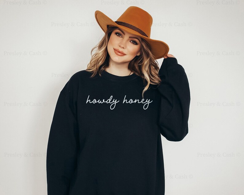 Pode incluir: Uma camisa de moletom preta com o texto "howdy honey" em escrita cursiva branca. A modelo est&aacute; usando um chap&eacute;u marrom.