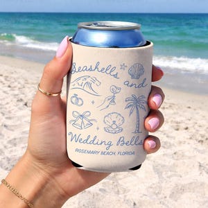 Cream can cooler held at the beach with “Seashells and Wedding Bells” printed in blue. Beachy bridal theme includes a shell, wave, palm tree, and rings. Customized with Rosemary Beach, Florida.