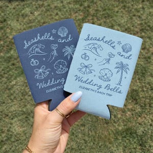 Two can coolers held in hand on grass, one navy and one light blue. Each features a “Seashells and Wedding Bells” design with palm tree, wave, and cocktail icons for a beach bachelorette.