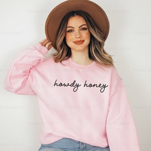Pode incluir: Uma camisola cor-de-rosa com o texto "howdy honey" em escrita cursiva preta. A modelo est&aacute; a usar um chap&eacute;u castanho e cal&ccedil;as de ganga azuis.