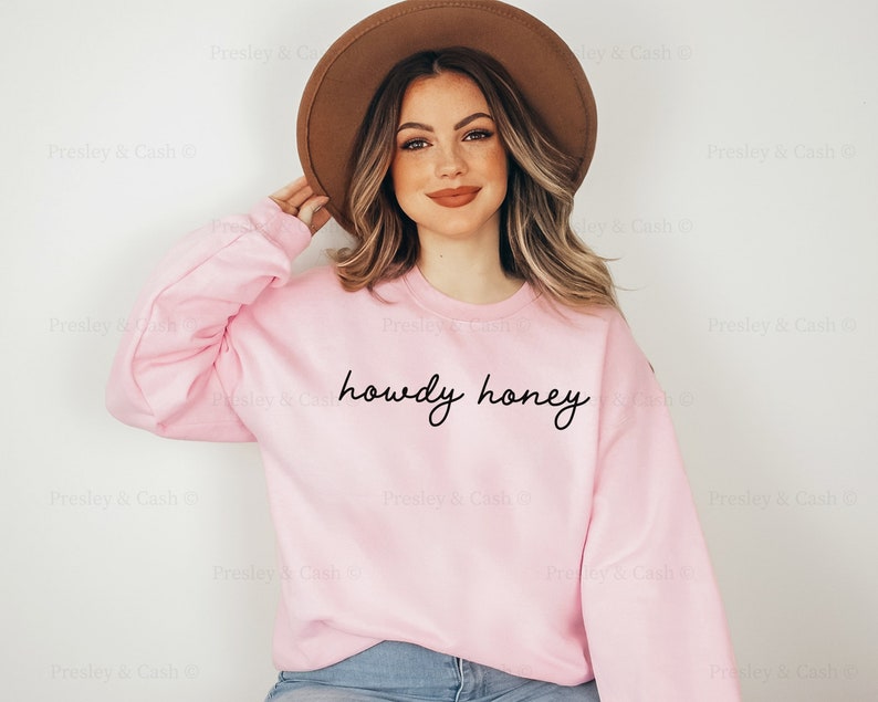 Pode incluir: Uma camisola cor-de-rosa com o texto "howdy honey" em escrita cursiva preta. A modelo est&aacute; a usar um chap&eacute;u castanho e cal&ccedil;as de ganga azuis.