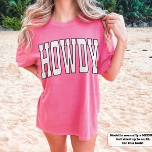 May include: Pink oversized t-shirt with the word "HOWDY" printed in white on the front. The model is wearing the shirt with the sleeves rolled up.