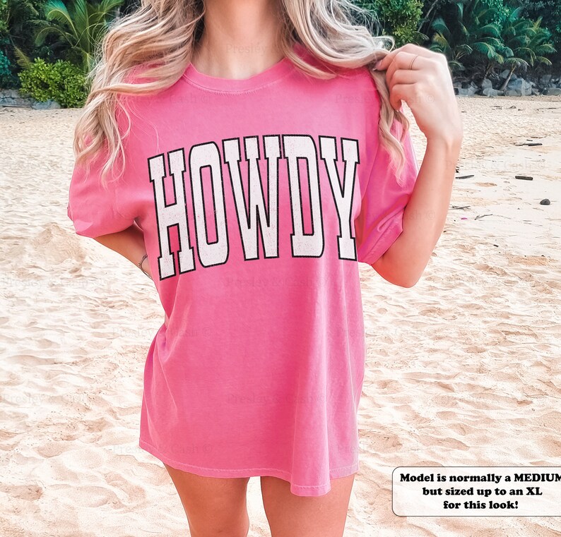May include: Pink oversized t-shirt with the word "HOWDY" printed in white on the front. The model is wearing the shirt with the sleeves rolled up.