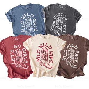 Flat lay of western bachelorette shirts in a variety of colors and sayings, including “Wild Wild Wife,” “Wild Wild Bach,” “Wild Wild Gals,” and “Let’s Go Girls.” Perfect for country-themed bachelorette parties or group gifts.