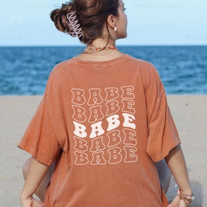 K&ouml;nnte beinhalten: Eine Frau tr&auml;gt ein rostfarbenes T-Shirt mit dem Wort "BABE", das in einer Retro-Schriftart auf dem R&uuml;cken wiederholt wird. Das Model sitzt auf einem Sandstrand mit dem Meer im Hintergrund.