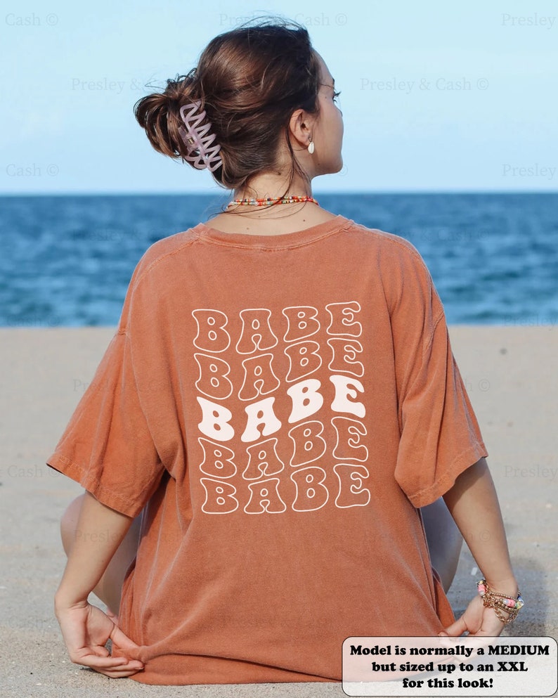 K&ouml;nnte beinhalten: Eine Frau tr&auml;gt ein rostfarbenes T-Shirt mit dem Wort "BABE", das in einer Retro-Schriftart auf dem R&uuml;cken wiederholt wird. Das Model sitzt auf einem Sandstrand mit dem Meer im Hintergrund.