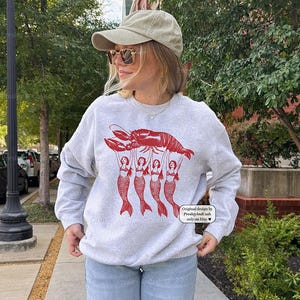 May include: A light gray sweatshirt featuring a red lobster graphic held up by five red mermaid figures. The text "Original design by Presley And Cash only on Etsy" is printed on the lower right corner.