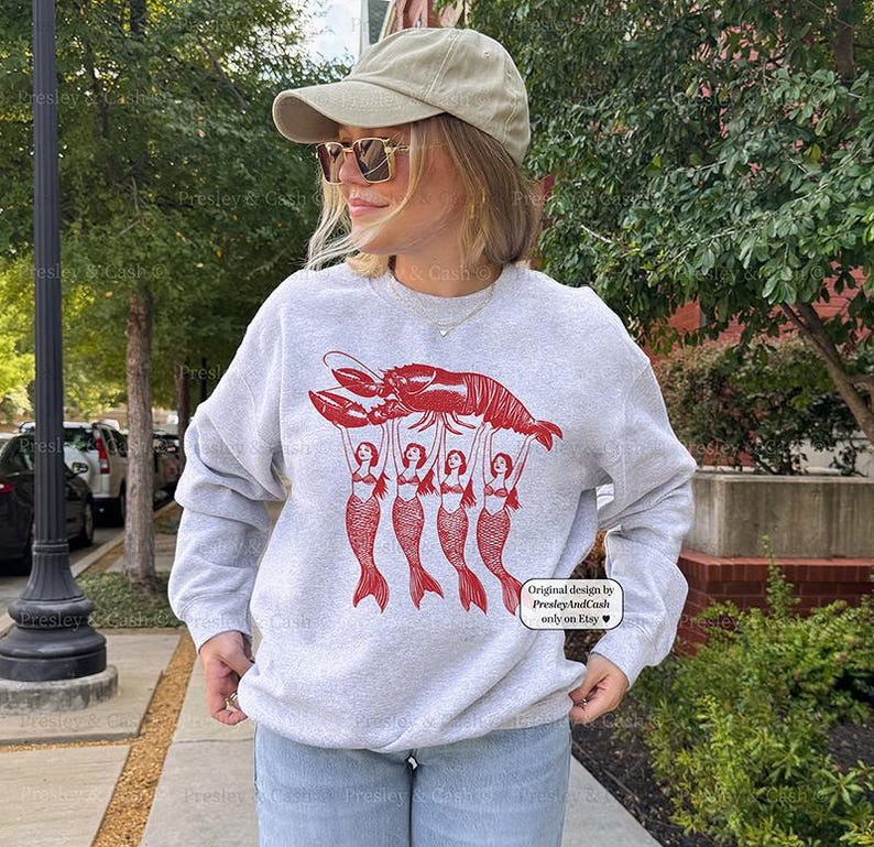 May include: A light gray sweatshirt featuring a red lobster graphic held up by five red mermaid figures. The text "Original design by Presley And Cash only on Etsy" is printed on the lower right corner.