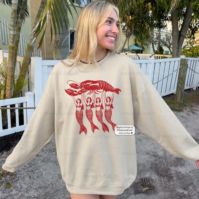 May include: A beige sweatshirt featuring a red graphic of a lobster held up by four mermaids. The text "Original design by PresleyAndCash only on Etsy" is printed on the lower right corner.