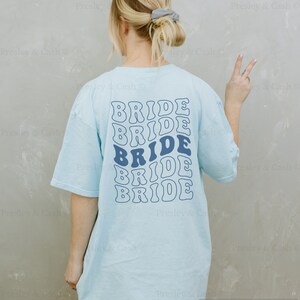 K&ouml;nnte beinhalten: Ein hellblaues T-Shirt mit dem Wort "BRIDE" f&uuml;nfmal in einer Retro-Wellenschrift wiederholt. Das Model tr&auml;gt Gr&ouml;&szlig;e XXL, tr&auml;gt aber normalerweise Gr&ouml;&szlig;e M.