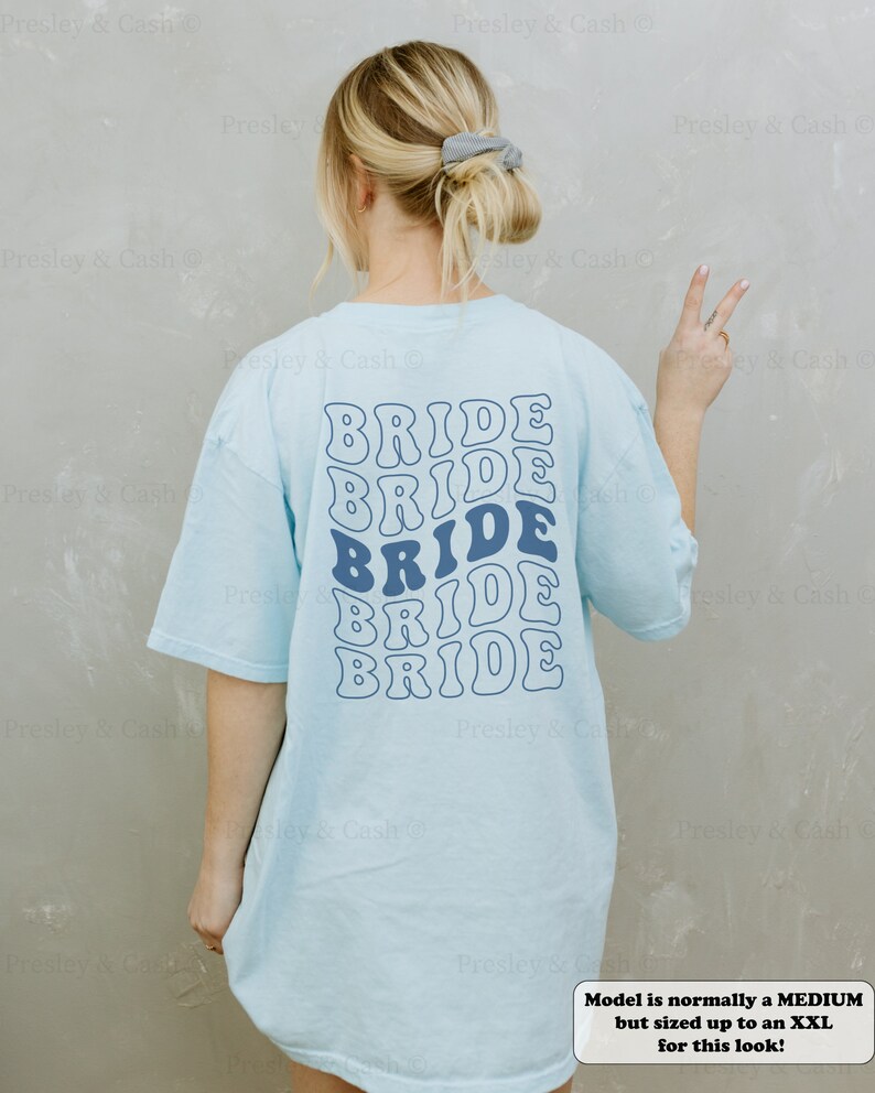 K&ouml;nnte beinhalten: Ein hellblaues T-Shirt mit dem Wort "BRIDE" f&uuml;nfmal in einer Retro-Wellenschrift wiederholt. Das Model tr&auml;gt Gr&ouml;&szlig;e XXL, tr&auml;gt aber normalerweise Gr&ouml;&szlig;e M.