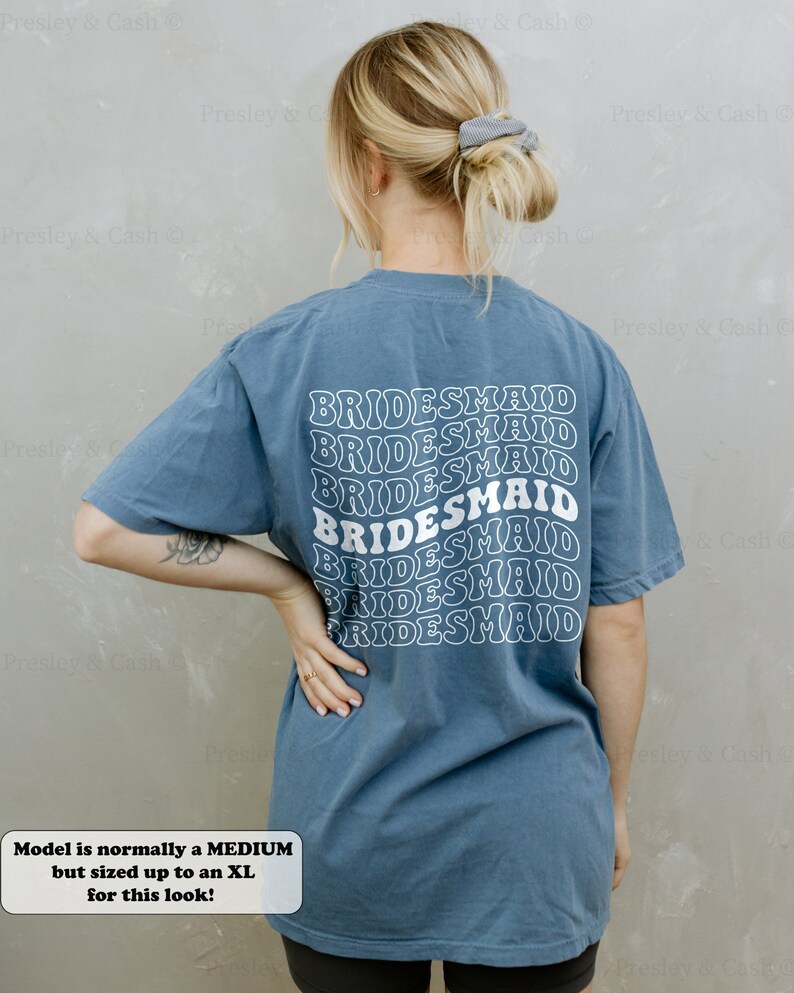 K&ouml;nnte beinhalten: Ein blaues T-Shirt mit dem Wort "BRIDESMAID" in einer Retro-Schriftart auf der R&uuml;ckseite. Das Model tr&auml;gt Gr&ouml;&szlig;e XL.