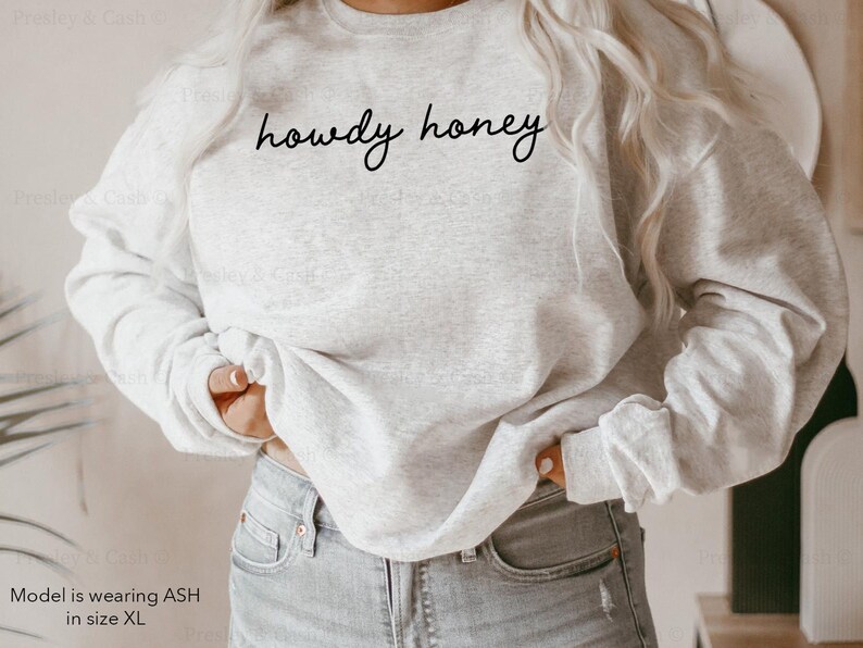 Pode incluir: Uma camisa de moletom cinza com o texto "howdy honey" em letras cursivas pretas.