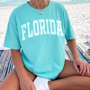 May include: A turquoise t-shirt with the word "FLORIDA" printed in white block letters. The shirt is worn with white sneakers. The person is sitting on a striped blanket on a beach.