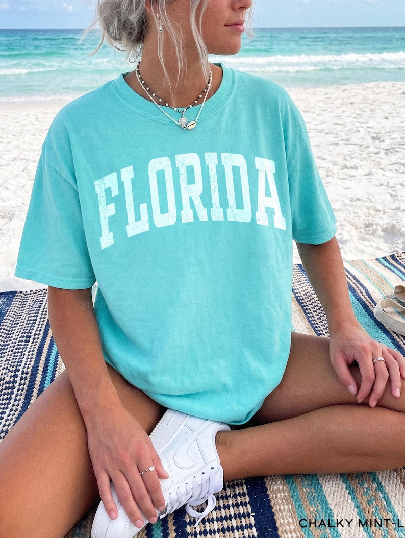 May include: A turquoise t-shirt with the word "FLORIDA" printed in white block letters. The shirt is worn with white sneakers. The person is sitting on a striped blanket on a beach.