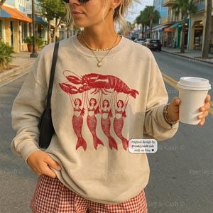 May include: Beige sweatshirt with a red lobster graphic held up by four mermaids. The text "Original design by PresleyAndCash only on Etsy" is on the lower right. The person is wearing red and white checkered shorts.