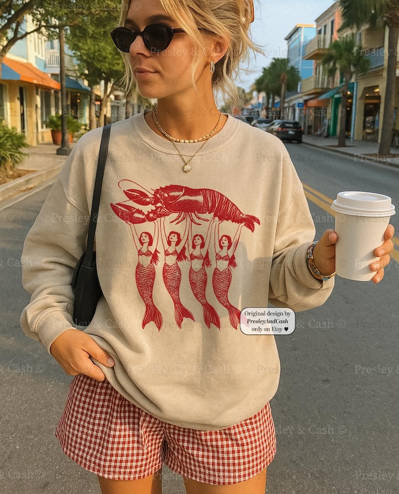 May include: Beige sweatshirt with a red lobster graphic held up by four mermaids. The text "Original design by PresleyAndCash only on Etsy" is on the lower right. The person is wearing red and white checkered shorts.