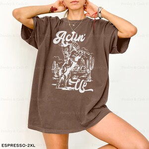 May include: A brown oversized t-shirt with a white graphic of a cowboy riding a bucking horse. The graphic also includes the text "Actin' Up".