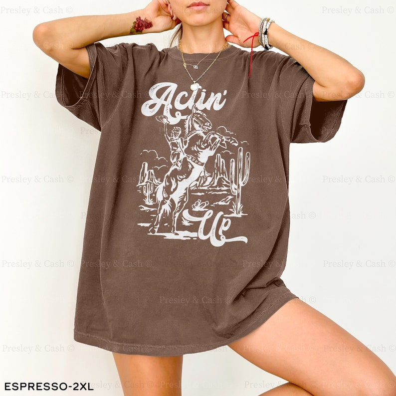 May include: A brown oversized t-shirt with a white graphic of a cowboy riding a bucking horse. The graphic also includes the text "Actin' Up".