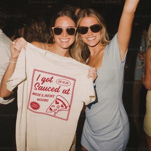 May include: Two smiling people wearing sunglasses hold up a cream-colored t-shirt with the text "I got Sauced at Maddie & Jason's Wedding" in red. The shirt also features a pizza slice graphic and the date 06-15-26.