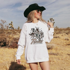 May include: White sweatshirt with a graphic of the state of Texas, a cowboy, and the text "Texas United States of America Since 1845." The sweatshirt is long-sleeved and has a relaxed fit. The image is set against a desert backdrop.