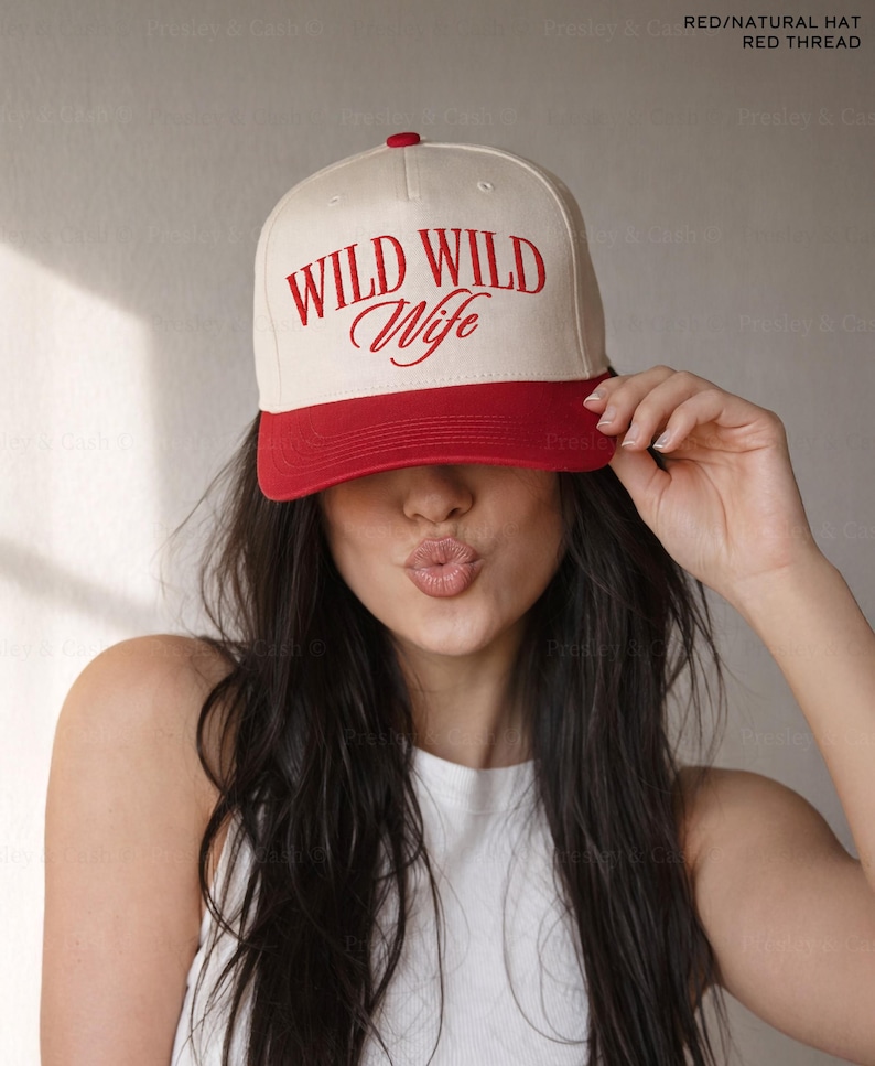 May include: A baseball cap with a cream-colored crown and a red brim. The cap features the words "WILD WILD Wife" in red script across the front. The hat is being held up by a person.
