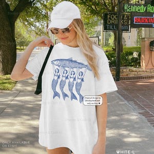 May include: White t-shirt featuring a blue illustration of a large fish held up by four mermaids. The shirt has the text "Original design by PresleyAndCash only on Etsy" and "ONLY AVAILABLE ON ETSY!"