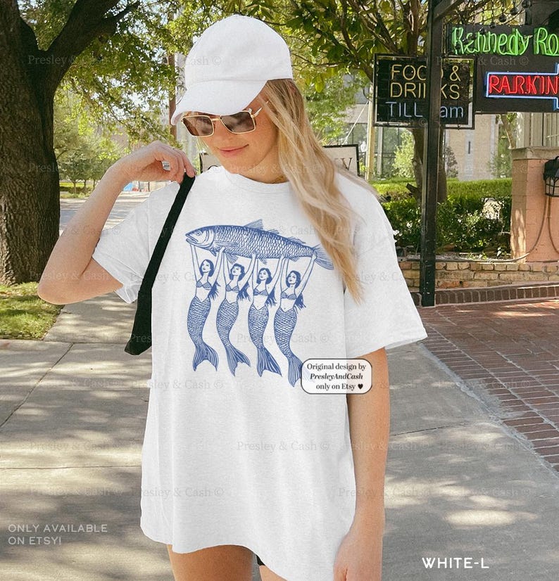 May include: White t-shirt featuring a blue illustration of a large fish held up by four mermaids. The shirt has the text "Original design by PresleyAndCash only on Etsy" and "ONLY AVAILABLE ON ETSY!"