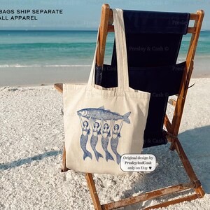 Peut inclure: Un sac fourre-tout en toile blanche avec une illustration bleue de quatre sir&egrave;nes tenant un poisson. Le texte "*TOTE BAGS SHIP SEPARATE FROM ALL APPAREL" est en haut du sac. Le sac est pos&eacute; sur une chaise de plage en bois sur une plage de sable blanc.
