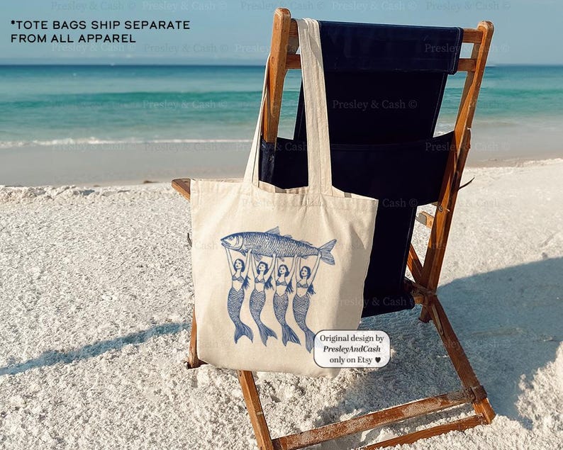 Peut inclure: Un sac fourre-tout en toile blanche avec une illustration bleue de quatre sir&egrave;nes tenant un poisson. Le texte "*TOTE BAGS SHIP SEPARATE FROM ALL APPAREL" est en haut du sac. Le sac est pos&eacute; sur une chaise de plage en bois sur une plage de sable blanc.