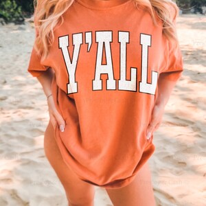May include: A woman wearing a burnt orange t-shirt with the text "Y'ALL" printed in white. The model is normally a medium size but is wearing a size XL.