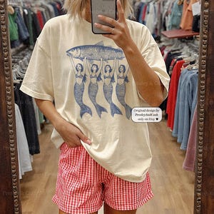 May include: Ivory-colored t-shirt featuring a blue illustration of a fish held up by four mermaids. The shirt is paired with red and white checkered shorts. Text on the shirt reads "Original design by PresleyAndCash only on Etsy".