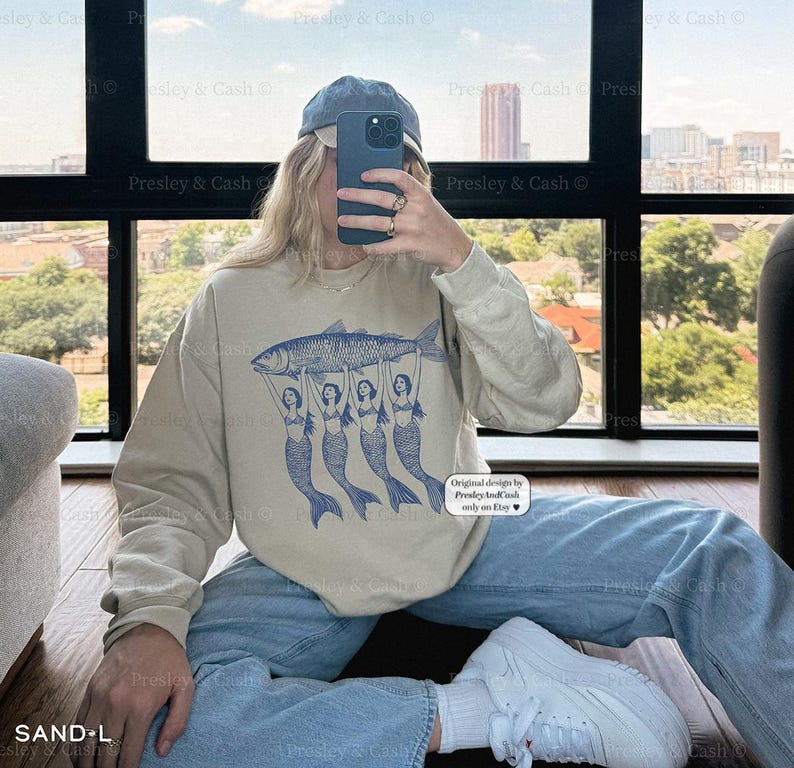 Peut inclure: Un sweat-shirt beige clair avec un imprim&eacute; graphique de quatre sir&egrave;nes tenant un poisson bleu. Le texte "Original design by Presley & Cash" est imprim&eacute; dans le coin inf&eacute;rieur droit du sweat-shirt.