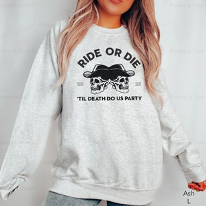 Peut inclure: Un sweat-shirt gris avec le texte "RIDE OR DIE" et deux cr&acirc;nes en graphiques portant des chapeaux de cowboy. Le texte "'TIL DEATH DO US PARTY" est sous les cr&acirc;nes. Le sweat-shirt est disponible en couleur cendr&eacute; et en taille L.
