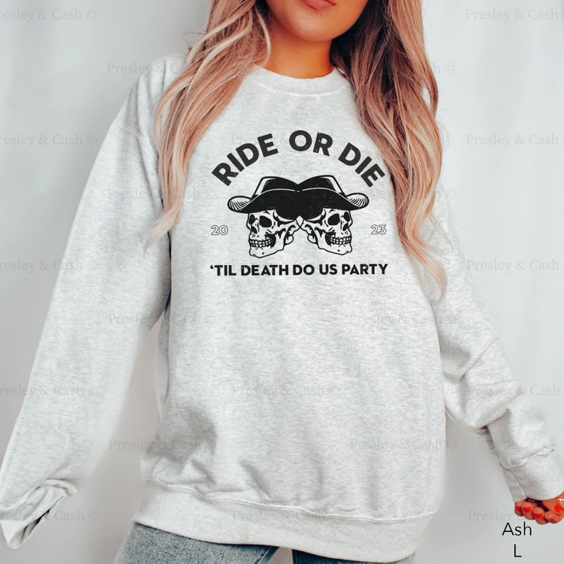 Peut inclure: Un sweat-shirt gris avec le texte "RIDE OR DIE" et deux cr&acirc;nes en graphiques portant des chapeaux de cowboy. Le texte "'TIL DEATH DO US PARTY" est sous les cr&acirc;nes. Le sweat-shirt est disponible en couleur cendr&eacute; et en taille L.
