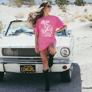 May include: A pink t-shirt with a white graphic of a cowboy riding a horse and the text "Actin' Up" on the front. The shirt is being worn by a person standing in front of a white car with a California license plate that reads "COWGIRL".