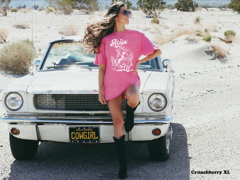 May include: A pink t-shirt with a white graphic of a cowboy riding a horse and the text "Actin' Up" on the front. The shirt is being worn by a person standing in front of a white car with a California license plate that reads "COWGIRL".