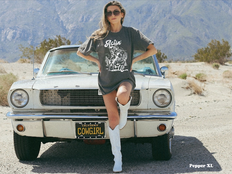 May include: A woman wearing a gray t-shirt with the text "Actin' Like" and a graphic of a cowboy riding a horse. She is standing in front of a white vintage car with a license plate that reads "California Cowgirl".