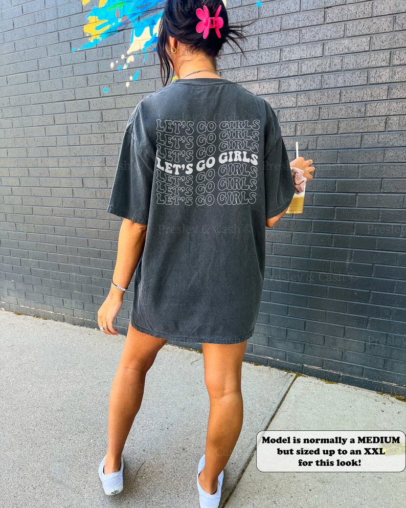K&ouml;nnte beinhalten: Ein schwarzes Oversize-T-Shirt mit dem Text "Let's Go Girls", der sechsmal in Wei&szlig; wiederholt wird. Das Model tr&auml;gt das Shirt und wei&szlig;e Sneaker. Der Text "Model is normally a MEDIUM but sized up to an XXL for this look!" ist am unteren Rand des Bildes sichtbar.