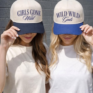 May include: Two baseball caps with contrasting brims. One is navy blue, the other royal blue. The caps have the phrases "GIRLS GONE Wild" and "WILD WILD Gals" embroidered in navy thread. The hats are cream-colored with a dark blue brim.